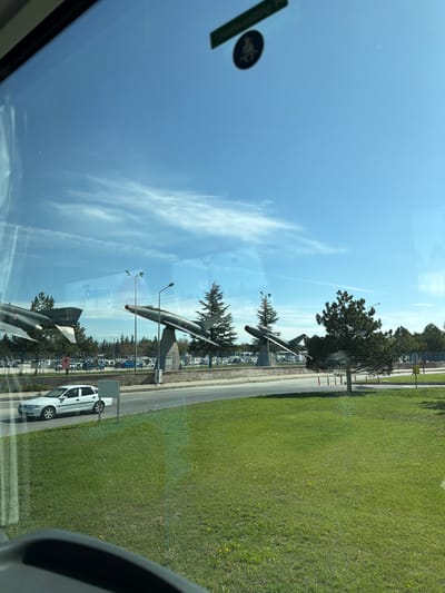 The image captures an outdoor scene from the interior of a vehicle, looking through a window. The primary subjects are three dark-colored jet aircraft, likely fighter jets, mounted on display poles. These aircraft are positioned in a row, parallel to a paved road or pathway. Behind the road, a wide expanse of neatly maintained green grass stretches towards the bottom of the frame, dotted with a few small yellow flowers, possibly dandelions.
Interspersed among the aircraft are several trees, predominantly evergreens. Tall light poles or flagpoles are also visible near the display. On the right side of the frame, a white car is parked or driving on the road. The sky is bright blue with scattered wispy white clouds, indicating clear and sunny daytime weather.
The view is partially obscured by reflections on the vehicle's window. A dark oval sticker with an indistinct light-colored symbol is visible in the top-left of the window reflection. Other subtle reflections, such as the curve of the vehicle's interior on the far right, also appear. The markings on the tail fin of the foreground aircraft appear to be an insignia, possibly resembling a national flag, consistent with the location context of Eskisehir, Turkey. The scene depicts a static aircraft display, potentially at a museum, a memorial, or an air force base entrance in Eskisehir.