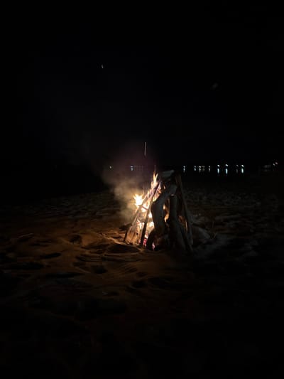 A nighttime scene features a crackling campfire against a dark, textured wall. The firelight illuminates the rough surface of the wall, revealing irregular patterns. Above the fire, a framework of branches is partially visible. Smoke rises from the flames, blurring the darkness beyond. Faint light from an unknown source appears in the upper section of the image, creating a subtle contrast against the dominant black backdrop.