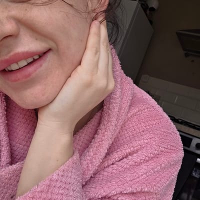 A close-up, tilted portrait features a person from the chin upwards, wearing a soft, pink fluffy robe. The person's left hand is gently resting along their jawline and chin. Their mouth is slightly open, revealing white teeth, and the lips have a reddish-pink hue, suggesting a natural lip color or product. Dark, slightly wavy hair is visible near the ear and neck on the right side of the frame. The skin shows natural texture with visible pores and minor blemishes.

The background is softly out of focus but shows hints of an indoor setting, possibly a home in Harrogate, United Kingdom. Elements such as a dark surface (potentially a countertop or table), white wall tiles, and what appears to be a mirror or frame can be discerned. The lighting is soft and ambient, characteristic of an indoor environment during the day. The overall scene conveys a sense of candidness and relaxation. No text is visible in the image.