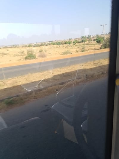 The image depicts a static view captured from inside a vehicle, looking outward through a window, located in Barakin Kuru III, Nigeria. The lower half of the frame is significantly occupied by reflections on the window glass, revealing parts of the vehicle's dark interior, specifically a headrest and seat structure on the lower right, and other indistinct dark surfaces. Several faint, curved scratch-like marks are present on the window glass. A dark, vertical vehicle pillar or frame element is visible along the far right edge, featuring a small, circular yellow object.

Outside the window, a two-lane asphalt road, dark grey in color, traverses horizontally across the mid-ground, flanked by unpaved, light brown shoulders. Beyond the road, the landscape extends as an arid, undulating terrain covered with dry, yellowish-brown soil and sparse, low-lying scrub vegetation. In the distant mid-ground and background, a line of green trees marks a slight rise in the topography, suggesting a more fertile area or a distant settlement. Several indistinct, light-colored structures are faintly visible among these trees. A tall utility pole with crossarms stands on the right side of the background, near the tree line, with a second, less distinct pole visible further to the left. The sky above is a clear, pale blue, transitioning to a lighter, hazy shade near the distant horizon. No people or active interactions are visible in the external scene.