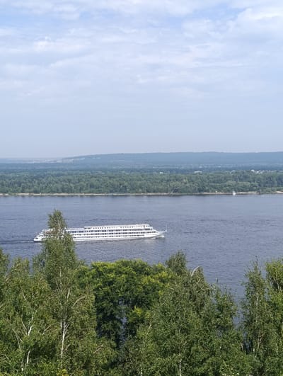 A long, white passenger ship with multiple decks is traveling on a wide, dark blue river. The ship has numerous windows and railings. In the foreground, the tops of lush green trees, including some with white bark, are visible, obscuring the lower portion of the image. Across the river, a dense, dark green forest covers the land, with rolling hills and a hazy sky in the distance. The sky is a pale blue with scattered white clouds. The location is Solnechnaya Polyana, Russia.