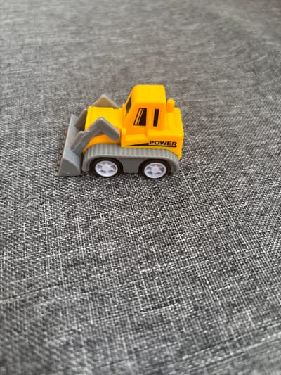 A yellow toy bulldozer sits on a textured gray fabric surface. The bulldozer has a gray blade at the front and black details on its cabin, including what appears to be the word "POWER" printed in black on the side. The wheels are white. The image is taken from a slightly elevated angle, with the bulldozer positioned in the center left of the frame. The fabric surface has a visible weave pattern. There is no indication of the city of Rodnichkovoye, Russia, in the image.