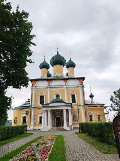 The photograph depicts the Resurrection Cathedral in the city of Uglich, Yaroslavl Oblast, Russia. The cathedral, a prominent example of 17th-century Russian church architecture, is painted a bright yellow with white detailing and features multiple green onion domes topped with golden spires. It is situated within a park-like setting, with a paved walkway leading up to its entrance. Flanking the walkway are meticulously maintained flower beds bursting with colorful blooms of pink, yellow, and white, interspersed with grey-green foliage. Tall green trees frame the left side of the image, and a dense green hedge runs along the right side of the walkway. The sky above is overcast with a pale, diffused light. There are no people or animals visible in the scene. The cathedral exhibits some scaffolding on its right side, suggesting ongoing restoration or maintenance work.