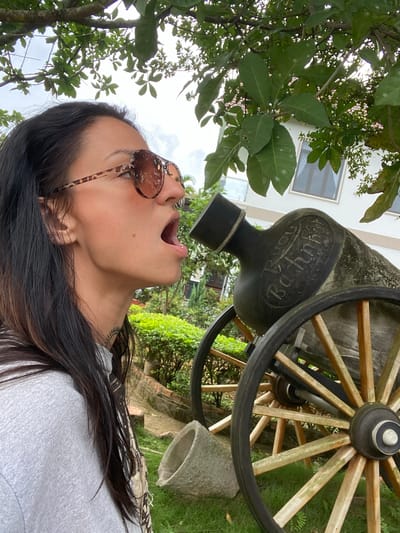 The media file shows a woman from the chest up, looking upwards with her mouth open. She is wearing sunglasses with a tortoiseshell pattern and a light gray shirt. Her long, dark hair is flowing. In the background, a large wooden cart wheel is visible, along with a stone object and some greenery. The scene appears to be outdoors, with trees and a building with windows in the distance. The lighting suggests it is daytime. The text "Pond Bachnhop" is visible on the side of the cart wheel. The location is identified as Thôn 2, Vietnam. The woman's pose and expression could convey surprise or playfulness.