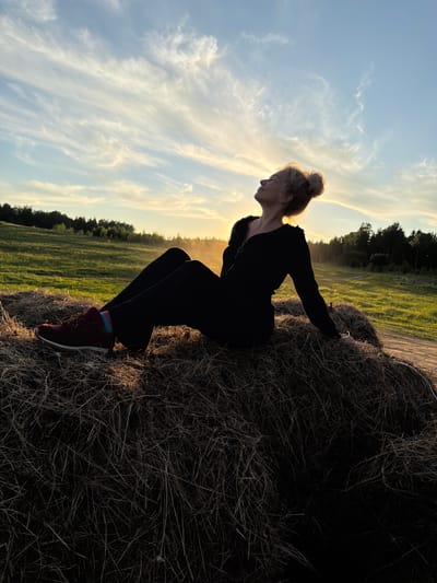 An outdoor shot captured during the golden hour, likely at sunset or sunrise, depicts a person silhouetted against a bright, cloudy sky and a sun-drenched field. The image is oriented with the horizon running diagonally from the top left to the bottom right.
The central subject is a person, appearing female, with hair styled in a bun. She is in a crouched or squatting position, leaning forward and resting her hands on a large, dark hay bale or stack of dried grass that occupies the right side of the frame. She wears dark, long-sleeved clothing and dark red sneakers with vibrant, multi-colored socks visible at the ankles. Her profile is visible, with her head tilted downwards. The bright light from behind illuminates the edges of her hair and body, creating a strong silhouette.
In the background, a vast, green grassy field extends towards a dark treeline under a bright sky. The sky is predominantly light blue with soft, wispy clouds stretching across it, glowing with warm yellow and orange hues near the horizon, indicating the low position of the sun. The scene suggests a peaceful, rural setting, potentially in the Аист Снт area of Russia, characterized by open fields and distant forest. No visible text is present.