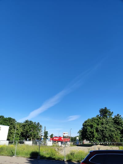 The image captures a bright, sunny day in Saue, Estonia. The sky is a vibrant, clear blue with a faint contrail stretching across it. In the foreground, a grey metal fence delineates an area of overgrown grass and yellow wildflowers. Beyond the fence, a road or pathway is visible, and further back, a large, lush green tree dominates the right side of the frame.
In the middle ground, there are several buildings. A white building is partially visible on the left, and to its right, a series of mature green trees line the horizon. A red truck with "Rimi" branding is prominent in the center, suggesting a commercial area. Another building with a sign that appears to read "MAPRI" is also visible. The general impression is one of an outdoor scene in a developed area, possibly a commercial district or a residential neighborhood with some construction or landscaping work underway, indicated by the temporary fencing. There are no people visible in the image. The overall lighting suggests it is daytime, likely late morning or early afternoon due to the clear sky and bright conditions.