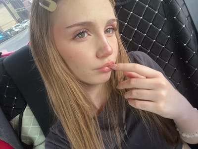 A young woman with long, light brown hair, a light complexion, and green eyes is captured in a close-up, selfie-style photograph. She is wearing a dark gray t-shirt and a bracelet of round, white beads on her left wrist. Her right hand is brought up to her lips, with her index finger touching her lower lip. She has a subtle makeup application, with noticeable mascara and blush. A beige hair clip is visible in her hair near the top of her head. The background suggests she is inside a vehicle, with a quilted black seat cover with white stitching visible behind her to the right. To the left of the frame, out of focus, is a street scene with several parked cars, suggesting an urban environment. The lighting indicates it is daytime, with natural light illuminating her face. The image is likely taken in the city of Novaya Otradovka, Russia, given the context of the request.