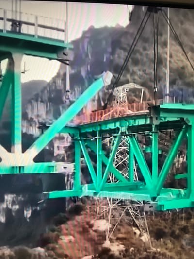 The image shows a construction scene, likely a bridge under construction, with large green metal structures dominating the foreground. In the background, a mountainous or hilly terrain is visible, with some vegetation. The lighting suggests it could be daytime. There are no people clearly visible. The scene appears to be an industrial or engineering project. There is no information in the image that can identify the location as Vangaži, Latvia.