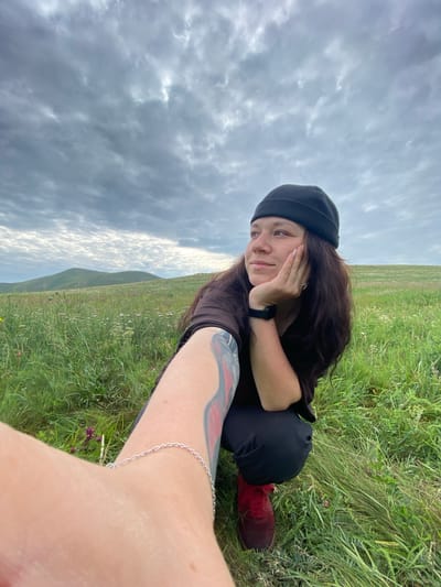 A person is crouching in a grassy field under a cloudy sky. The person is wearing a black beanie, a dark t-shirt, and black pants. They have long, dark hair and are resting their chin on their hand, looking to the right. A tattoo is visible on their left arm. A silver bracelet is on their left wrist. Their left foot, wearing a red shoe, is visible in the foreground. The field is covered in green grass and scattered wildflowers. In the background, rolling hills are visible under a dramatic, overcast sky. The location is in the vicinity of Абзаново, Russia.