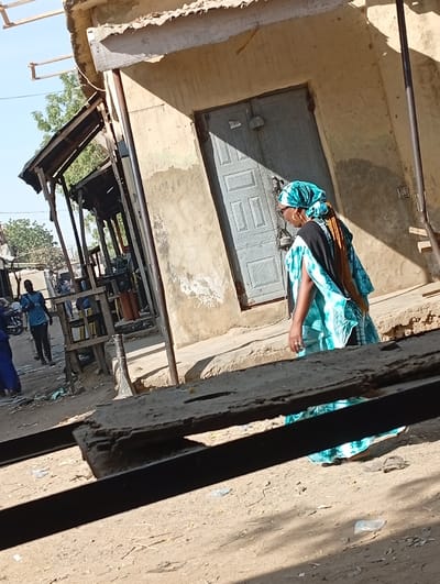 The image captures an outdoor street scene in Kofar Ruwa, Nigeria, under bright sunlight. A woman in the mid-ground walks left, wearing a teal patterned headwrap, a matching long dress with black sleeves, gold hoop earrings, eyeglasses, and a ring on her left hand. Her hair is styled in reddish-brown braids. To her right stands a light brown plastered building with a closed, grey metal double door featuring a geometric relief pattern. A distinct shadow falls diagonally across this door. In the background on the left, several rudimentary wooden stalls with flat roofs are visible, lining an unpaved dirt path where multiple individuals are present, including one person in a blue shirt. Scattered plastic debris litters the dry ground. Green trees are visible against a clear blue sky in the far background. A dark, out-of-focus wooden beam partially obstructs the lower foreground horizontally.