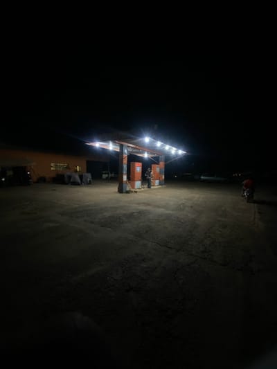 The image captures a nighttime scene at a gas station in Igarra, Nigeria. A central, orange-and-grey structure with a metal canopy is brightly illuminated by multiple overhead lights, casting light onto two visible fuel pumps. An individual in dark clothing stands adjacent to the rightmost fuel pump. To the right of the pumps, a person wearing a red garment is seated on a motorcycle, facing away from the viewer.

To the left of the fuel pumps, an orange-colored, single-story building with at least one illuminated window is present. In front of this building, several objects are visible under dark coverings. A light-colored vehicle, possibly a van, is partially visible near the building's right side. The ground surface of the station appears to be unpaved dirt or gravel, showing various textures and minor cracking, particularly towards the foreground. The overall environment outside the immediate light cast by the canopy and building is enveloped in deep darkness.