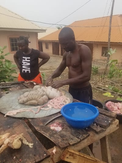 The scene takes place in Pati Ridge, Nigeria, at the coordinates 7.81 latitude and 6.704 longitude. A man is preparing meat on a table in a residential backyard. He is shirtless and wearing dark pants. He is using a large knife to cut and prepare meat. There are various cuts of meat on the table including large sections of intestines, and a blue basin of water. A woman is standing behind the man wearing a black shirt that says “HAVE” on it, with orange shorts and a dark colored headscarf. There is another building behind the man, with an orange corrugated metal roof. The buildings are in various states of construction and have exposed brick walls. The ground is dirt and some areas are overgrown with weeds. There is a small tree behind the woman.  The weather is overcast.