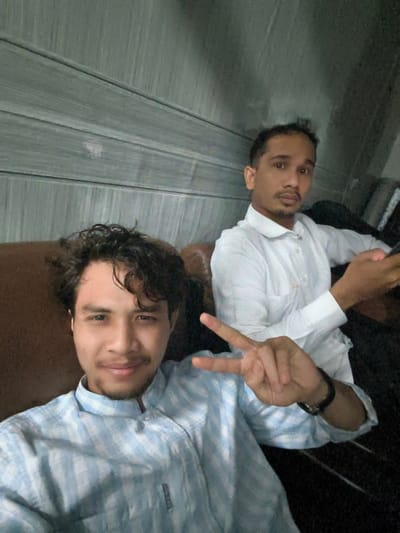 The image shows two men sitting on a brown couch. The man in the foreground is wearing a light blue and white checkered shirt and making a peace sign with his right hand. He has curly dark hair and a mustache. The man in the background is wearing a white shirt and holding a smartphone in his right hand. He has short dark hair and a goatee. The background appears to be a wall with horizontal wooden paneling. The scene is likely indoors, possibly in a residential setting in Pulodolang, Indonesia.