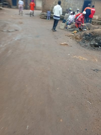 The image depicts a dusty, unpaved street scene in a settlement, likely in Dong, Nigeria, characterized by its earthy tones and arid conditions. The environment is predominantly brown and granular, with some areas showing patches of light-colored dust or soil. In the background, simple, block-style buildings with muted colors suggest a rural or semi-urban setting.
Several individuals are present in the scene. On the left side, two figures stand facing away from the camera, one in a white top and patterned skirt, and the other in a bright orange shirt. Further in the background, a group of people are gathered. One woman, wearing a red outfit, is actively engaged in cooking over an open fire. She is holding a utensil and appears to be stirring something in a pot. Several other individuals, including children, are seated or standing around this cooking area. The clothing of the people varies, with some wearing traditional attire and others more contemporary garments.
Objects visible include a white bucket, various bowls, and the cooking fire itself. There are also some larger rocks or stones scattered around the area, one of which appears to be the base for the cooking setup. The ground surface is uneven and shows signs of wear and tear, consistent with a well-trafficked unpaved road. The overall impression is of a communal gathering and daily life unfolding in an outdoor setting.