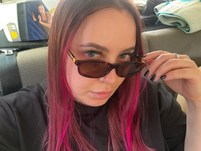 A young woman with long, pink-streaked brown hair, wearing sunglasses and a black shirt, is looking at the camera. She is sitting in a car, as indicated by the car seat visible in the background and a product box with an image of a woman sleeping on an air mattress. A folded blue and white towel is also visible in the background. The woman has a nose ring and is wearing a ring on her left hand. Her fingernails are painted black.
