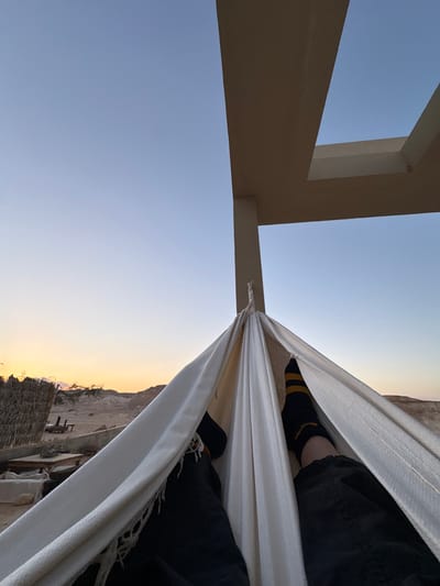 The image captures a first-person perspective from within a white fabric hammock, showing the lower legs of an individual wearing black trousers and black socks with yellow stripes. The hammock is suspended from a beige, modern architectural structure featuring a vertical column and an L-shaped overhead beam. Beyond the hammock and structure, a desert landscape unfolds under a twilight sky, transitioning from light blue at the top to orange and yellow near the horizon, indicating either sunrise or sunset. In the left midground, a structure made of dried straw or thatch is partially visible, alongside what appears to be outdoor furniture (tables and chairs) on sandy ground. Distant light-colored hills or mountains define the horizon. The scene is located in the Arava Tichona Regional Council, Israel.