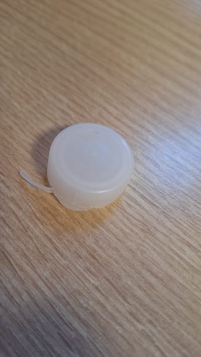 A white plastic bottle cap rests on a light brown wooden surface. The cap is round with a ribbed edge and a small, broken piece of plastic extending from its side. The wooden surface has a distinct grain pattern running diagonally from the top left to the bottom right of the image. The lighting suggests it could be daytime, with soft illumination casting a slight sheen on the wood. There is no visible text in the image. The scene is simple and focuses on the solitary object against a textured background.