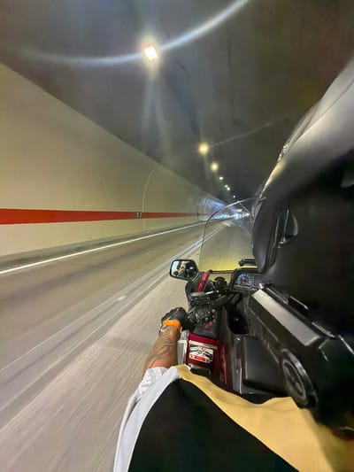 A first-person perspective shot from a motorcycle as it travels through a tunnel. The tunnel walls are a light beige color, with a prominent horizontal red stripe running along the left side. The tunnel ceiling is illuminated by multiple lights, some of which are visible as bright circles due to the camera's focus. The road surface is paved and appears to have a white line marking. The motorcycle's handlebars, dashboard with a speedometer, and a portion of the rider's helmet are visible in the foreground. The rider's left arm, wearing an orange glove, is also visible, along with tattoos. A side mirror on the motorcycle reflects a blurred image of the tunnel interior and possibly another rider. The tunnel appears to be located in the city of Đurmani, Montenegro, as indicated by the reference to the location.