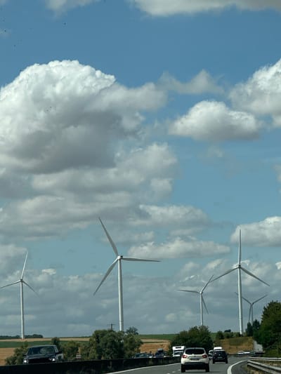 This is an outdoor, eye-level, vertical, daytime photograph taken from a vehicle on a highway. The scene depicts a landscape with multiple wind turbines in various stages of rotation against a cloudy blue sky. The foreground shows a highway with several cars and trucks, including a white SUV in the center lane, a black SUV on the left, and a truck on the right. The middle ground features rolling hills with fields of dry grass and scattered trees. The background is dominated by the imposing structures of the wind turbines, with the sky above filled with puffy white and grey clouds. The location is identified as Nouvion-le-Comte, France.