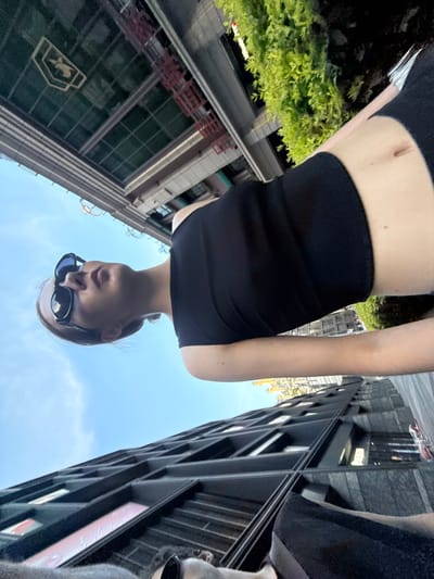 The image captures a unique low-angle view of a person in an urban setting, oriented as if looking upwards at the subject against the sky. The subject is a young individual, likely female, with light-colored hair, wearing a black crop top and black bottoms, with her bare midriff visible. She is wearing dark sunglasses, obscuring her eyes, and her face is partially visible as she looks down towards the camera or slightly away.

The scene is dominated by a bright blue sky with some scattered white clouds, indicating clear daytime weather. Surrounding the subject are tall, modern city buildings constructed with glass and dark paneling, typical of a bustling urban environment. A notable detail is a building on the viewer's left (when the image is correctly oriented) that prominently features the golden logo of the Bank of China and the Chinese characters "中国银行" (Bank of China) on its facade, strongly suggesting the location is China. Green foliage from trees or bushes is also visible near this building, adding a touch of nature to the concrete jungle. On the right, another dark, tall building shows faint, unreadable text or lighting within its windows.

The image has a casual, perhaps selfie-like, quality, with the low camera angle creating a dramatic perspective of the person against the towering cityscape. There is no specific activity or event taking place other than the person being present and photographed in this urban landscape.