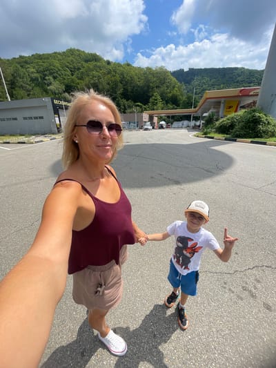 An adult female and a young male are holding hands and taking a selfie at a gas station with a mountainous backdrop. The female, in the foreground, wears a burgundy tank top, beige shorts, white sneakers, and sunglasses. She has blonde hair and is smiling. The male, to her right, wears a white t-shirt with cartoon characters, blue shorts, a baseball cap, and sneakers. He is also smiling and making a rock-on hand gesture. They are on an asphalt surface, with a gas station canopy, buildings, and lush green mountains visible in the background. The scene appears to be in Agoy, Russia, suggested by Cyrillic signage on one of the buildings.