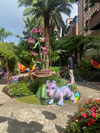 The image depicts a garden scene in Gramado, Brazil. A whimsical, brightly colored statue of a tiger lies on a green, circular base surrounded by flowers. The ground is paved with interlocking rectangular stones. Behind the tiger statue, there's a large, artificial plant with large, purple, flower-like structures. A man wearing a black jacket and khaki pants is standing in the background near a cluster of ferns. In the background, there is building with a Bavarian-style architecture and the sky. The entire scene is vibrant and colorful.