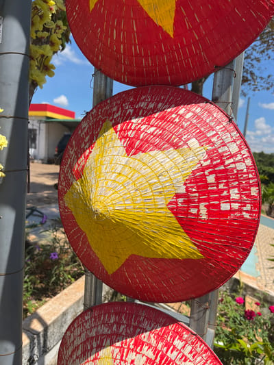 The image displays a close-up view of two conical hats, traditionally Vietnamese, painted with a red and yellow star pattern. These hats are suspended vertically on a metal pole, with one hat partially obscuring the other. The hats appear weathered, with chipped paint and visible straw weave. In the background, a building with a red and yellow roof is visible, along with lush green foliage and a bright blue sky with scattered clouds. A gray pole with some yellow flowers is positioned to the left of the hats. The scene is outdoors, suggesting a public space or roadside. The location of Ia Hru, Vietnam is not explicitly identifiable from the visual information provided in the image itself.