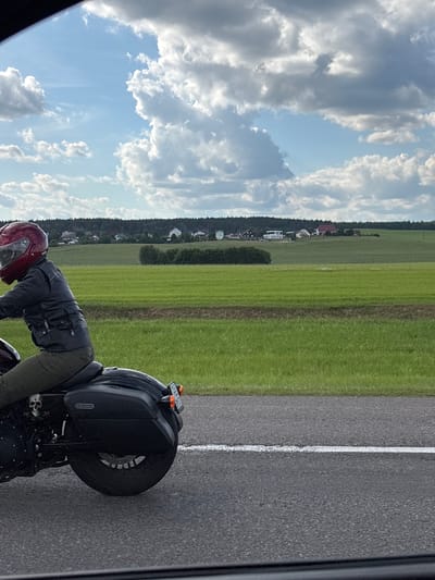 This is a vertical, slightly blurry, candid photograph taken from a low angle. The main subject is a person wearing a dark leather jacket, green pants, and a red helmet, leaning over the handlebars of a black motorcycle. The motorcycle is parked on the side of an asphalt road with a white line marking the lane. The road runs parallel to a vibrant green field, which is dotted with trees and a small village in the distance. The sky is filled with dramatic, white cumulus clouds against a bright blue backdrop.
The scene appears to be set in a rural location in Belarus, given the context. The sunlight suggests it is daytime, likely late afternoon or early evening due to the soft lighting and the angle of the sun, although this is not definitively clear. The weather appears to be pleasant, with a mix of sun and clouds. The person's posture suggests they might be resting or preparing to set off on their journey. The overall mood is one of a solitary ride through the countryside. There is no visible text in the image.