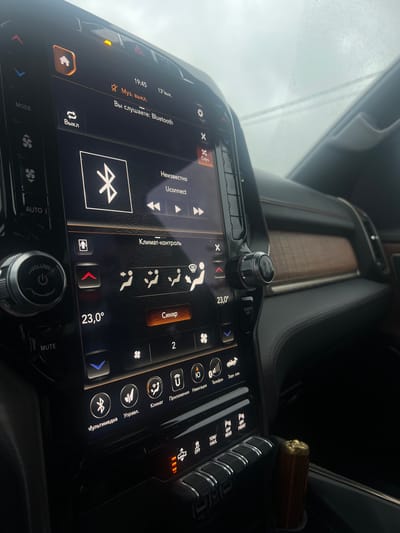 The image shows the interior of a car, focusing on the central touchscreen display and dashboard. The screen displays the time as 19:45 and indicates that music is off, with the text "Вы слушаете: Bluetooth" (You are listening: Bluetooth). A Bluetooth symbol is prominently displayed, along with "Неизвестно Uconnect" (Unknown Uconnect). The climate control settings are visible, showing a temperature of 23.0 degrees Celsius and icons related to air distribution. Below the climate control, there are icons for multimedia, control, climate, applications, navigation, phone, and "Зад. кам." (Rear camera). Several buttons and a rotary dial are also visible on the dashboard. The car's gear shifter, made of wood and metal, is also in view. The overall environment appears to be a car interior, with the background suggesting a cloudy or overcast day outside, possibly during rain due to some streaks on the windshield. There is no visible indication of the car's location being in Jõhvi, Estonia.