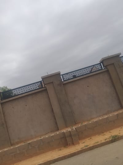 This image shows a wall with a decorative metal railing on top, situated in Gindi Nassarawa Kyarmi Galbi, Nigeria. The wall appears to be made of concrete blocks covered in a light brown stucco. There are two prominent pillars on the right side of the frame, also made of concrete with stucco. The metal railing features a geometric pattern in black. A few scattered patches of greenery are visible on the ground near the base of the wall, which is bordered by a concrete curb and a sandy area. The sky above is overcast and grey.