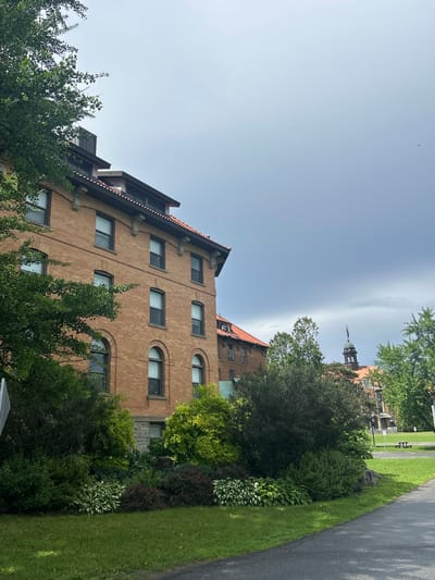 The image depicts a view of the exterior of buildings at McGill University's Macdonald Campus in Sainte-Anne-de-Bellevue, Canada. The foreground shows a paved pathway on the right, with a well-maintained lawn and a variety of green foliage, including trees and bushes, to the left. In the mid-ground, a prominent brick building with a tiled roof is visible, featuring numerous windows. Further in the background, other buildings with similar architectural styles can be observed, including one with a distinctive tower topped with a flag. The sky is overcast with heavy clouds, suggesting a possibly stormy weather condition. The overall scene conveys a campus environment with a mix of historic architecture and natural landscaping.