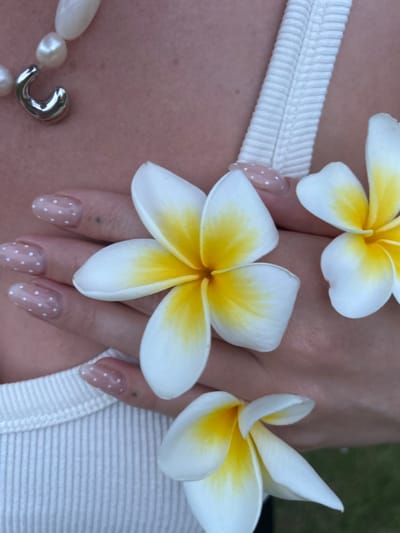 The scene is located in Corrimal, Australia. A close-up view presents the upper torso and left hand of an individual. The individual is wearing a white, ribbed tank top and a necklace featuring irregular white pearls and a silver, C-shaped pendant positioned on the left side of the neck/collarbone area. The left hand, positioned across the chest, displays nails manicured with a translucent or pale polish adorned with white polka dots. Three plumeria (frangipani) flowers, characterized by white petals and yellow centers, are present. One flower rests on the palm of the left hand. Another flower is partially visible in the upper right quadrant, resting on the skin. A third, partially obscured flower is visible at the bottom of the frame, resting on the white ribbed top. The background is out of focus, revealing a blurred green area consistent with outdoor vegetation or grass.