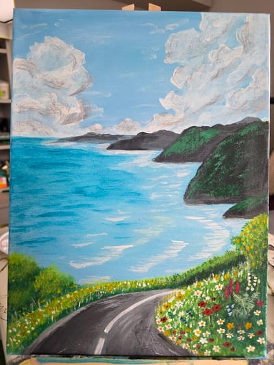 This is an acrylic painting on canvas depicting a scenic coastal road. The scene features a winding asphalt road in the foreground, bordered by lush green foliage and a vibrant array of wildflowers in white, yellow, red, and pink. Beyond the roadside, a bright turquoise ocean stretches out, meeting dark, verdant hills and mountains in the middle ground. In the background, more distant, muted hills are visible under a clear blue sky populated by fluffy white clouds. There are no people or specific landmarks to indicate a location within Claremorris, Ireland.