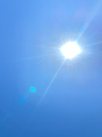 The image displays a clear blue sky with the sun prominently positioned in the upper right quadrant. The sun is very bright, emitting rays of light that extend outwards. There is a lens flare, appearing as a circular, cyan-colored spot, in the lower left quadrant of the image. A fainter, elongated lens flare is also visible near the cyan spot. The image appears to be taken outdoors on a sunny day. No specific location in the city of Police, Poland, can be determined from the image content.
