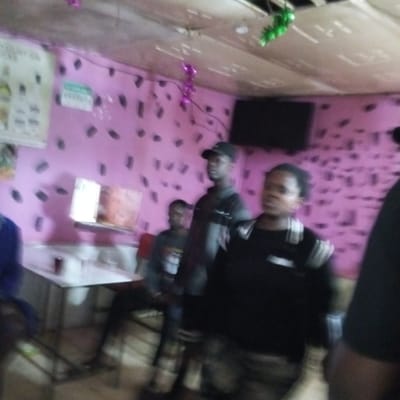The image shows an interior space in Kinoo ward, Kenya. The walls are painted pink with black irregular shapes, and the ceiling is a light, possibly fabric-covered, surface. A black TV is mounted on the wall. A white table with what appears to be glasses is visible, and a calendar or menu is on the wall near it. Several individuals are present, including one wearing a black baseball cap. Some are standing, while at least one person is seated. One individual wears a black sweater with white accents, while another has on a grey jacket over a dark shirt. Decorations, including green and purple streamers, are attached to the ceiling.