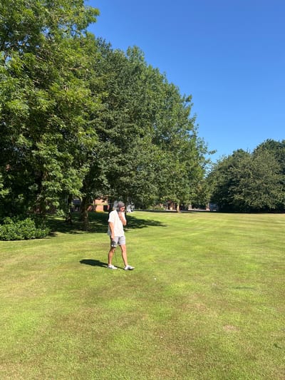 The image depicts a single male subject on a golf course under a clear blue sky, likely at Hornsea Golf Club in Hornsea, United Kingdom. The male, with fair skin and dark hair partially covered by a backward-facing dark baseball cap, is positioned slightly left of center, facing towards the right foreground. He wears a white short-sleeved t-shirt, grey shorts, white socks, and white athletic shoes with black stripes (Adidas). A dark object, possibly headphones, is visible around his neck or resting near his left ear. His left hand is raised near his mouth or chin, while his right hand holds a golf club vertically, resting its head on the green grass near his right foot. A white golf ball is visible on the grass directly in front of and slightly to the right of his feet. The ground consists of well-maintained green grass, characteristic of a golf course. Numerous mature deciduous trees with dense green foliage line the left side and background, casting distinct shadows, indicating bright sunlight. In the mid-ground, behind the trees, brick buildings with traditional architecture are partially visible. No other individuals or active play are observed.