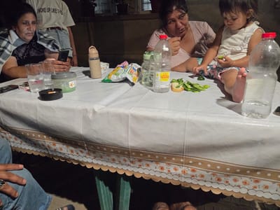 In Todor Ikonomovo, Bulgaria, an outdoor evening gathering takes place around a white plastic-covered table. Three individuals are visible: a woman on the left, partially obscured by a plaid blanket, holds a mobile phone. Another woman in the center, wearing a striped shirt, gestures with her hand. A young child sits to the right, wearing a white top and floral shorts, and appears to be eating. The table is laden with various items, including drinks in plastic bottles and glasses, a bag of snacks, an ashtray with a cigarette, and a container with a green substance. The table's edge is decorated with a floral pattern. The scene suggests a casual family meal or social gathering outdoors.