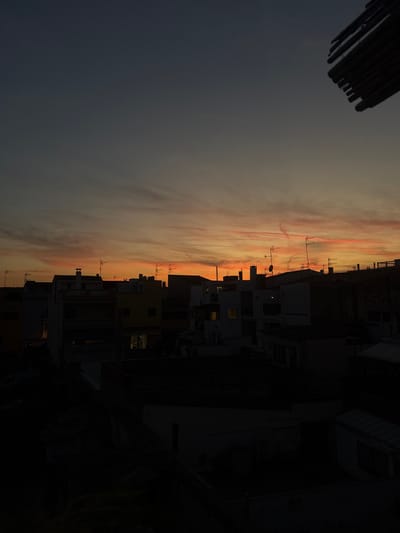 A low-angle, wide shot captures a dense urban skyline at dusk. The sky is a gradient of deep blue at the top, transitioning through orange and pink hues towards the horizon, indicative of a sunset. Wispy clouds are streaked across the sky, catching the fading light.
The cityscape is dominated by buildings of varying heights and architectural styles, silhouetted against the colorful sky. Many buildings have flat roofs, and several are adorned with numerous television antennas. Chimneys are also visible on some of the rooftops. Windows on some of the structures are illuminated, suggesting indoor activity.
In the foreground and lower portion of the image, the buildings are darker, almost entirely in shadow, obscuring finer details. The overall impression is of a residential or mixed-use urban area. The image appears to have been taken from a higher vantage point, such as a rooftop or balcony, looking out over the city.
