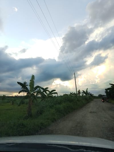 An eye-level, medium shot captures a rural road in Santa Cruz, Philippines, during what appears to be late afternoon or early evening. The sky is a dramatic mix of dark, heavy clouds and bright, sunlit patches. Power lines are visible stretching across the sky, supported by a wooden pole on the right side of the frame. To the left, lush green fields are dotted with banana trees and other vegetation. A rough, unpaved dirt road curves into the distance on the right, leading towards a small, motorized vehicle with passengers. The overall impression is one of a natural landscape under a dynamic sky.