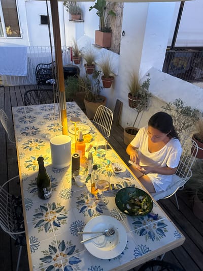 A woman sits at a table laden with food and drinks on a tiled patio in Vilanova i la Geltrú, Spain. She is wearing a white t-shirt and appears to be rolling a cigarette. The table is set with plates, cutlery, glasses, bottles of wine and orange juice, a salt shaker, a pepper mill, and a green salad bowl. In the background, a large umbrella shades the patio, and potted plants are visible on shelves and the ground. The scene suggests an evening gathering or meal.