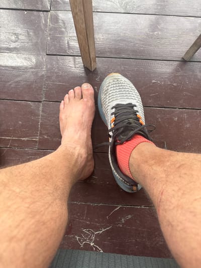 A person's bare left foot and right foot wearing a grey and white athletic shoe with orange accents and a red sock are positioned on a dark wooden surface. The wooden surface appears to be deck boards. A portion of a wooden chair leg is visible in the upper right quadrant of the image. The scene is outdoors, possibly in a rustic setting. There are no visible indications of the city of Arkhyz, Russia.