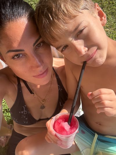 In this close-up, vertically oriented image, a child and an adult share a frozen drink on a grassy area, likely outdoors given the bright sunlight casting shadows and highlighting the textures of the grass. The subjects are positioned intimately, with the child's head resting on the adult's chest or shoulder. The child, positioned closer to the viewer, has fair hair and is looking upwards, with a black straw extending from their mouth to a clear plastic cup filled with a pink, icy beverage. The adult, appearing to be female with dark hair, is looking towards the camera with a gentle expression, her face partially obscured by the child. She is wearing a black bikini top adorned with a white celestial pattern and multiple gold necklaces. The child is wearing patterned swim trunks. The lighting suggests it is a sunny day, likely mid-day, and the overall atmosphere appears relaxed and joyful, hinting at a moment of shared enjoyment, perhaps during a summer outing. The location context indicates Beit Yitzhak-Sha'ar Hefer, Israel, suggesting a warm climate. There is no visible text in the image.