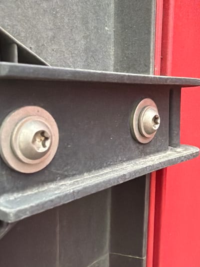 The image shows a close-up, eye-level view of two silver, hex-socket, cap screws with washers inserted into a textured, dark gray plastic bracket. The bracket is positioned horizontally, with a ribbed edge running along the bottom. To the right of the bracket, a bright red surface is visible, creating a stark contrast with the gray plastic. The lighting appears to be natural, casting soft shadows. The image does not contain any discernible location markers for Murfreesboro, United States of America, and there are no people or actions depicted.