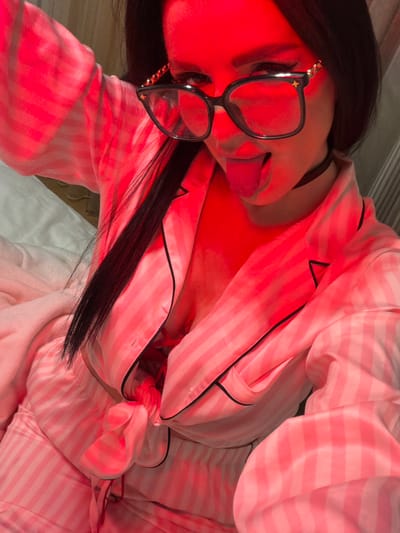 A person with long dark hair, wearing black glasses with gold accents and a black choker, is reclining in bed. They are wearing pink and white striped pajamas with black piping. The room is illuminated by red light.
