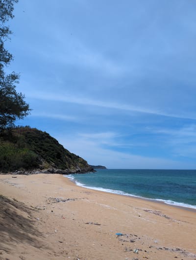 The image depicts a scenic view of a sandy beach in Sa Huynh Ward, Vietnam, stretching towards a rugged, tree-covered hillside on the left. The ocean, with its shades of blue and green, meets the shore with gentle waves. The sky is a clear blue, interrupted by thin, wispy clouds. The beach is mostly deserted, with some scattered debris, including plastic waste, visible on the sand. A lone, small boat is resting on the sand near the water's edge. The overall atmosphere is tranquil and natural, with a hint of human impact due to the presence of litter.