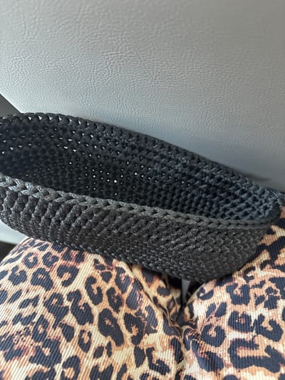 A black crocheted bag rests on a person's lap, who is wearing leopard-print pants. The bag is open, with its interior and rim visible. Behind the bag, a textured, light-colored surface, possibly a car seat or similar upholstery, is present. The background is out of focus, but the scene appears to be indoors or within a vehicle. There is no visible indication of the location being in Мастоцкі сельскі Савет, Belarus.