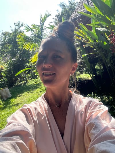 A woman with a blonde bun hairstyle smiles at the camera. She is wearing a light pink robe and two delicate gold necklaces. The woman is positioned outdoors, with lush green foliage and a thatched-roof structure visible in the background. The scene suggests a tropical or subtropical environment, possibly a resort or a private residence with extensive gardens. The sunlight indicates it is daytime. The location is Tambon Khlong Thom Nuea, Thailand.