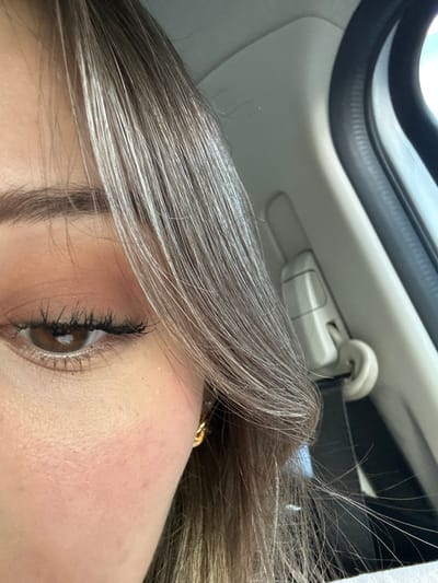 The image shows a close-up of a young woman's face. Her right eye is visible, with brown iris and black eyeliner. Her eyebrow is slightly arched, and she is wearing light-colored eyeshadow. Her skin appears pale with a light blush, and her long, straight, light brown hair is parted to the side, covering part of her face.  She is wearing a gold hoop earring in her right ear. The background suggests she is sitting in the passenger seat of a vehicle, with a part of the door, the window, and a seatbelt visible. The coordinates provided locate the scene in San Nicolás de los Garza, Mexico.