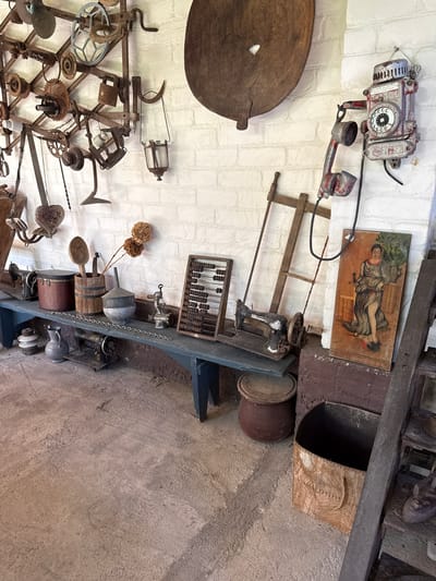 The image depicts an indoor scene featuring a collection of antique items displayed against a white brick wall and on a blue wooden bench. The objects appear to be stored in what could be a museum or a historical display area.
Dominating the upper left of the image is a complex arrangement of rusted metal gears, chains, and tools, possibly part of a historical machinery exhibit. To the right, a large, weathered wooden bowl or platter hangs on the wall. Below that, a vintage red and silver wall-mounted telephone with a rotary dial is prominent.
On the blue bench, various items are arranged. From left to right, these include what appear to be antique sewing machines, a dark cylindrical container, a wooden bucket holding utensils like a large spoon, a metal funnel, and dried flowers. Further to the right on the bench is a wooden abacus.
Below the bench, on the concrete floor, are more objects, including a metallic jug, a smaller sewing machine, and a large, dark-colored pot. To the right of the bench, leaning against the wall, is a painting of a woman in classical attire holding a sword over a prostrate figure. A wooden ladder-like structure is also visible on the far right. A woven basket sits on the floor near the ladder.
The lighting suggests an indoor environment, possibly a dimly lit room or a space with ambient light. The overall impression is one of a curated collection of historical artifacts.
There is no visible indication within the image to suggest this scene is located in Alagir, Russia. The description is limited to the visual elements present in the photograph.