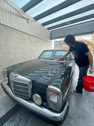 The scene depicts a male individual washing a dark green vintage Mercedes-Benz sedan inside a self-service car wash bay in Oberhausen, Germany. The man, with dark, wavy hair, is wearing a black t-shirt with a white graphic, blue jeans, and dark sneakers. He is positioned on the passenger side of the vehicle, actively scrubbing the car's hood near the windshield with a red rectangular sponge held in his right hand. His left hand holds a red plastic bucket containing water and suds. The Mercedes-Benz, featuring a prominent chrome grille, bumper, and auxiliary lights, is covered in white soap suds and water droplets. The car wash bay features light grey tiled walls on the lower section, corrugated metal siding above, and a translucent corrugated roof supported by dark beams. The concrete floor is wet and includes a metal drainage grate in the foreground. Outside the bay, a light blue/grey two-story building and bare trees are visible under an overcast sky.
